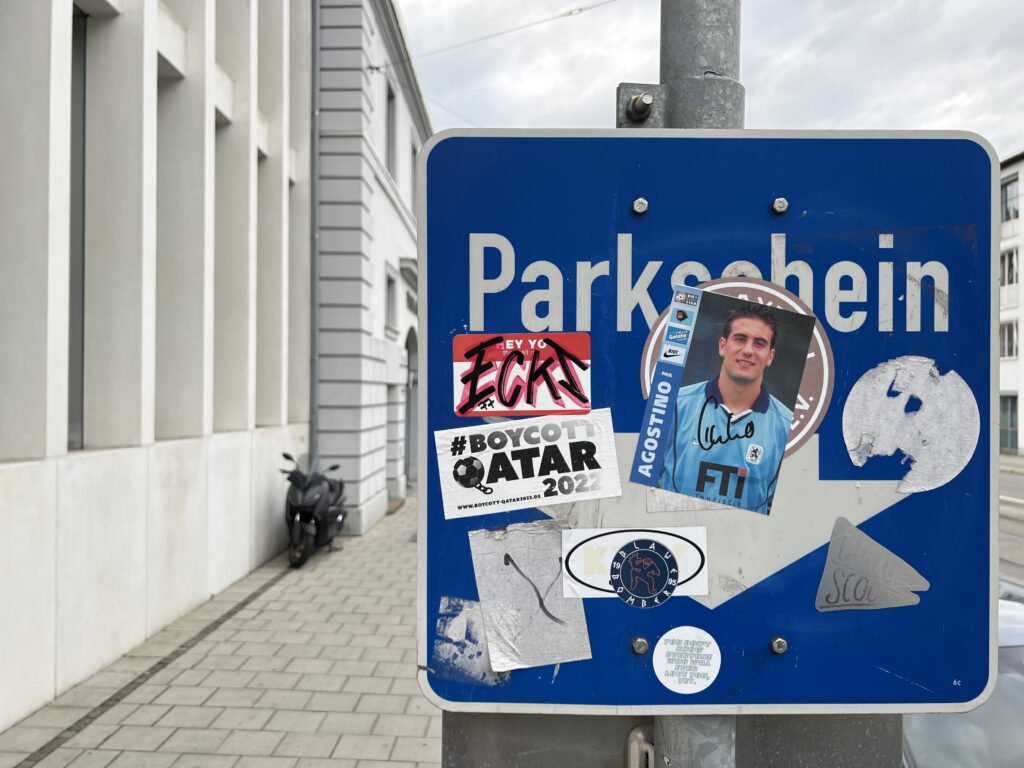 A street sign called "parkschein", which means "parking ticket". it is a photograph and in the background there is a big concrete building visible. on the street sign, lots of stickers can be found, so that the sign is barely readable anymore. The stickers are from several football fan clubs and initiatives, some of them are even layered over each other or scratched out, that shows a kind of remote rivality. 