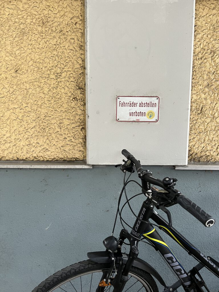 a photograph of a house-wall that has a sign "fahrräder abstellen verboten" which means "no bike parking here". the sign holds a small sticker for a bike demonstration event. furtheremore, direcly in front of the sign, a montain bike was parked.