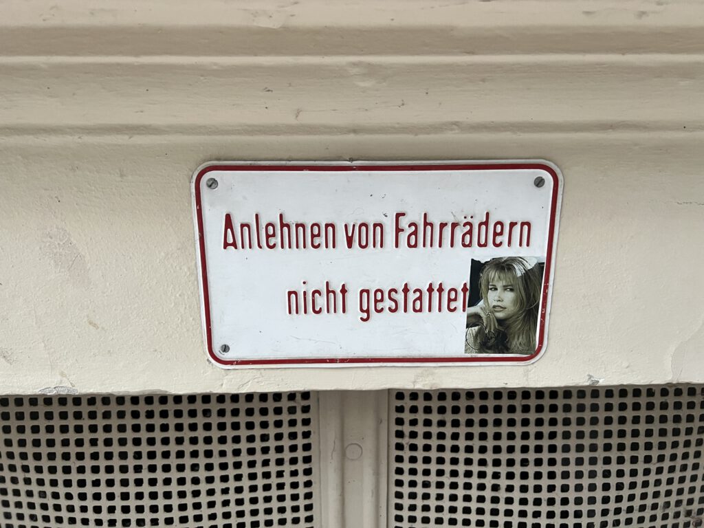 A photograph of the wall of a house with a sign "Anlehnen von Fahrrädern nicht gestattet" which means "Leaning bikes prohibited". There is a sticker on the sign that shows an former super-model. It turned out to be Claudia Schiffer.